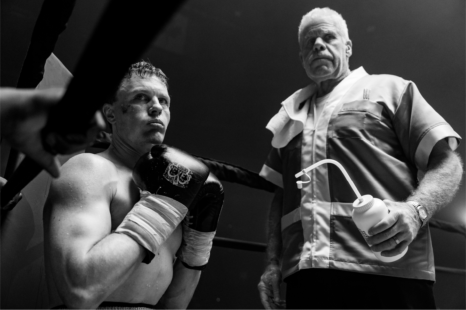Michael Pitt, nel ruolo di Mike Flannigan, siede concentrato e determinato sul ring, con i guantoni pronti per il combattimento, mentre Ron Perlman, nei panni di Stevie, il suo allenatore, lo osserva con intensità. La scena, in bianco e nero, cattura un momento di pausa e strategia durante il match, simbolo del legame tra il pugile e il suo mentore in Il Giorno dell’Incontro.