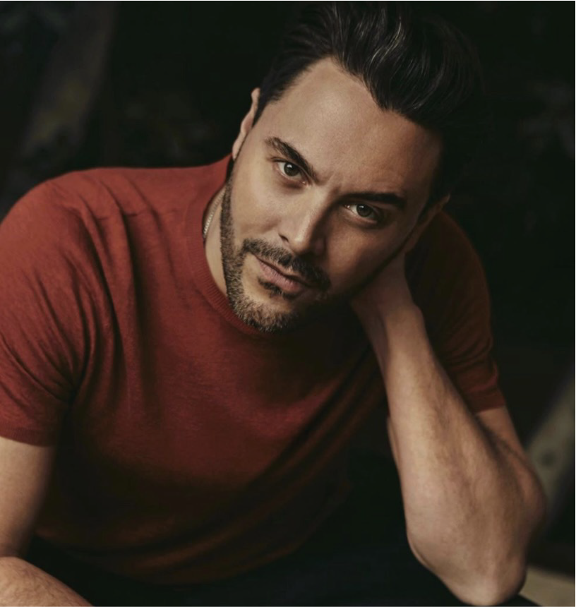 Jack Huston, regista di Il Giorno dell'Incontro, in un ritratto promozionale. Indossa una maglietta rossa e guarda intensamente verso l'obiettivo, appoggiando una mano al collo. La foto riflette la sua personalità creativa e la sua dedizione al cinema.