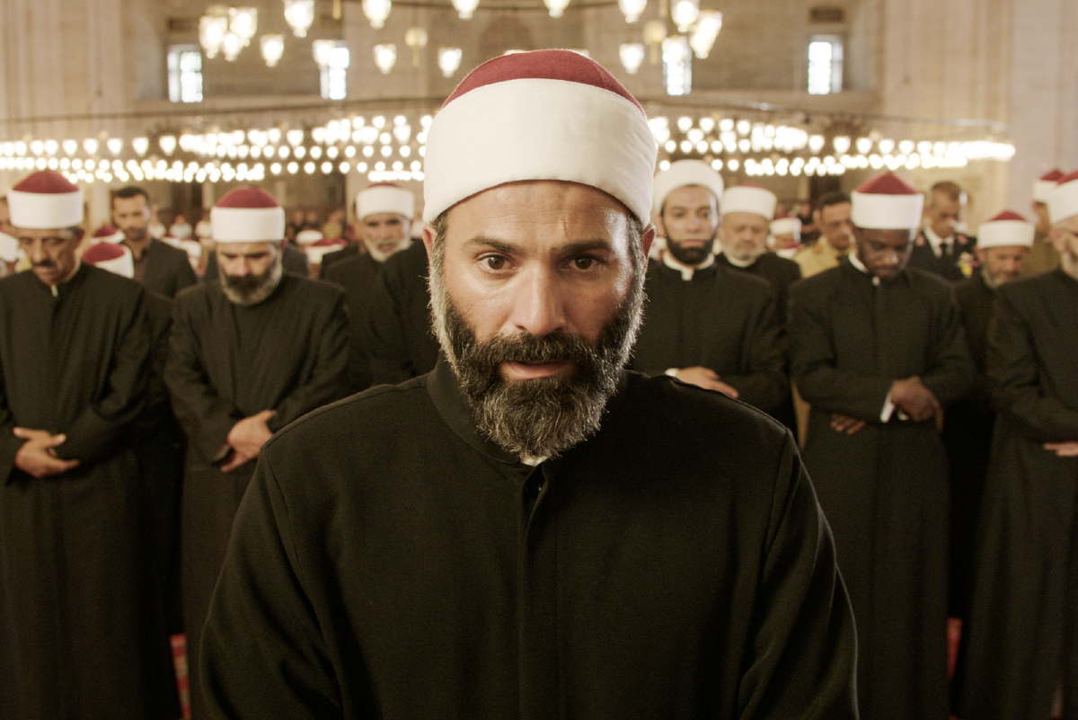 Un imam con barba nera e sguardo intenso guida la preghiera, circondato da decine di religiosi con turbanti.