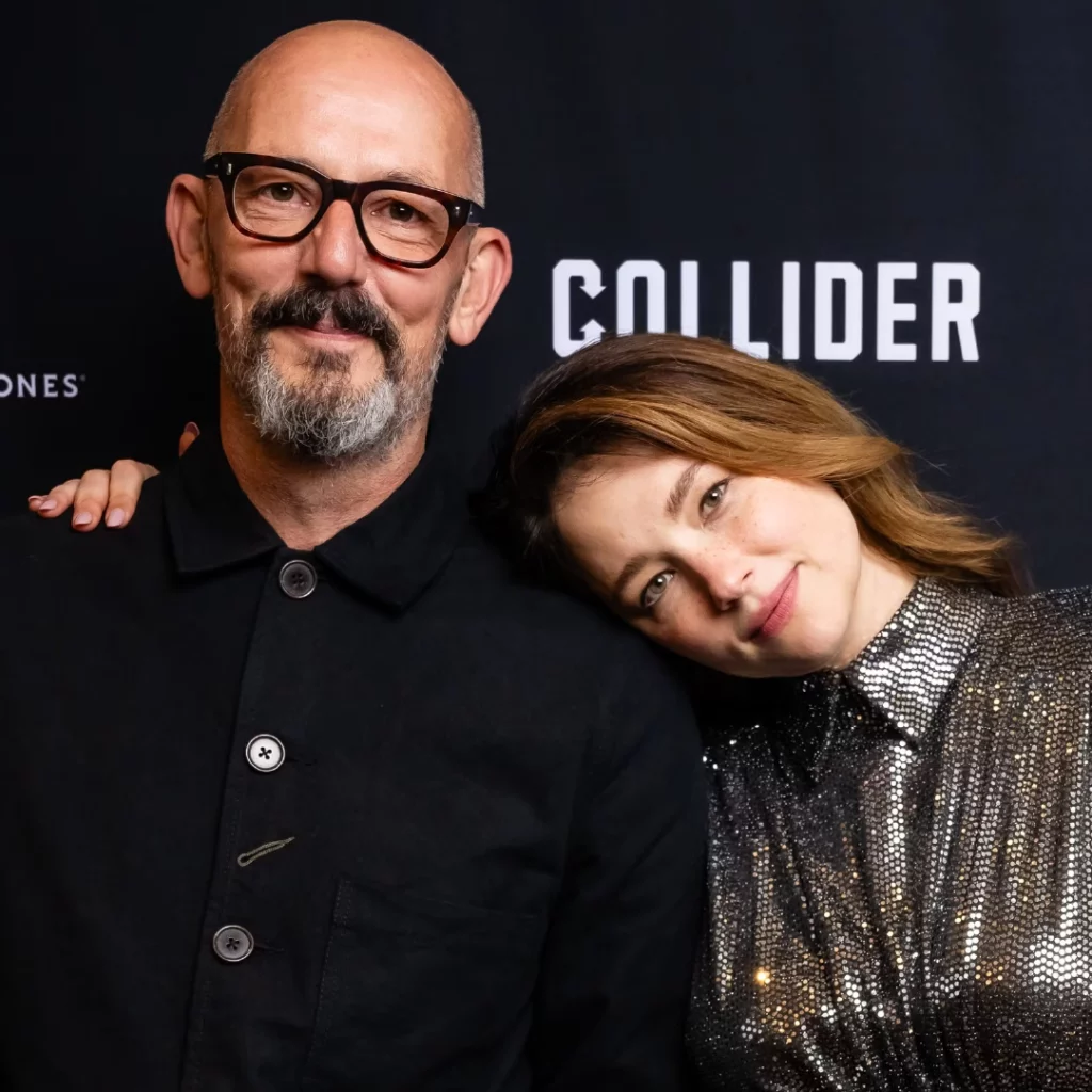 Thomas Napper, regista di Madame Clicquot, e Haley Bennett, protagonista del film, posano insieme sorridenti su un tappeto rosso con il logo di Collider sullo sfondo.