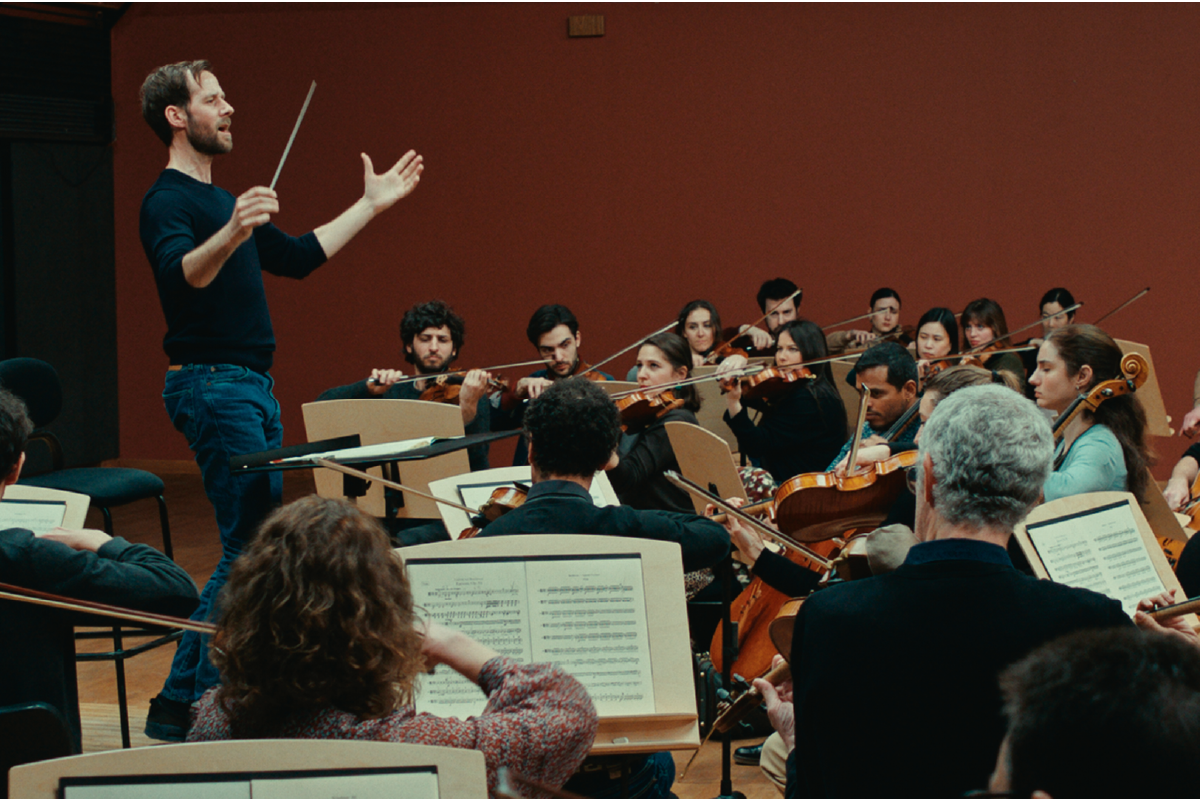 Scena dal film L&apos;Orchestra Stonata: un direttore d’orchestra con bacchetta alza le braccia mentre guida un gruppo di musicisti intenti a suonare strumenti ad arco in una sala prove, creando un’atmosfera intensa e coinvolgente.