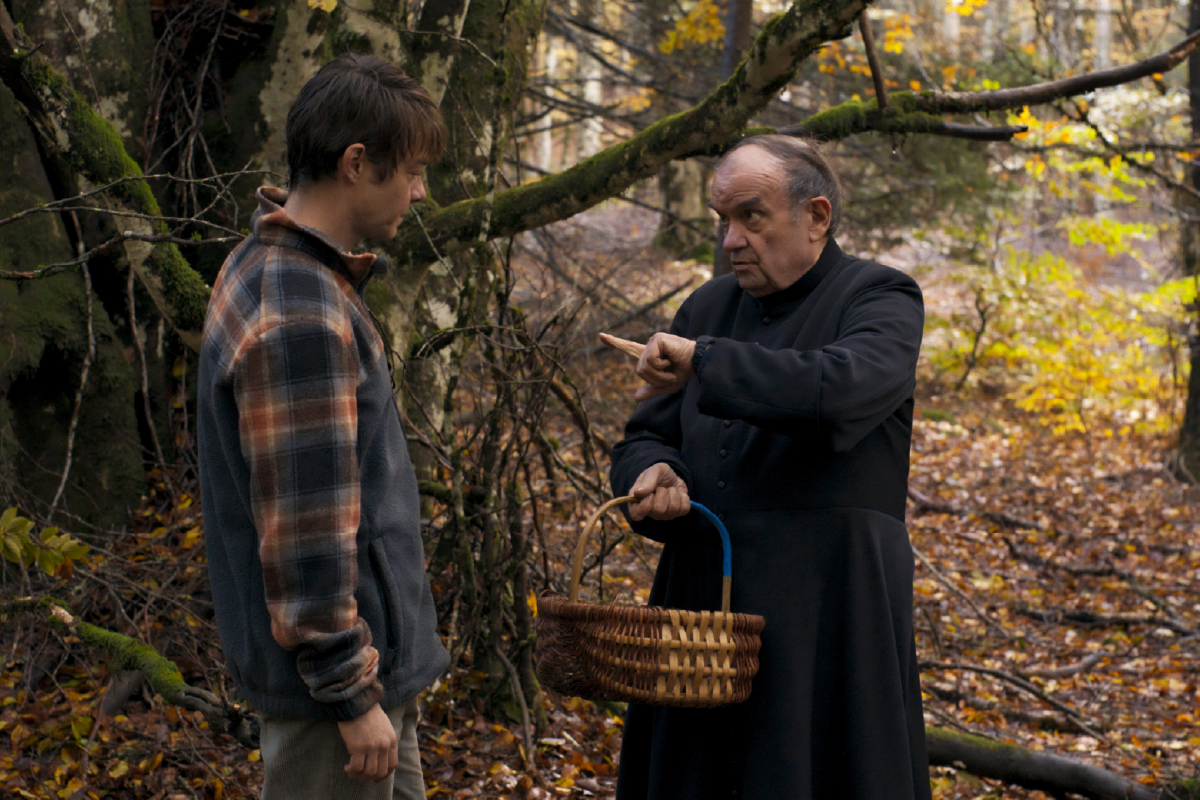 Scena tratta dal film “L’Uomo nel Bosco”. Jérémie (Félix Kysyl) si trova faccia a faccia con il prete (Jacques Develay) in una foresta autunnale. Il prete, con un’espressione severa, indica qualcosa con il dito mentre tiene in mano un cesto di vimini. Jérémie, in una giacca a quadri, ascolta attentamente. Sullo sfondo, alberi e foglie gialle creano un’atmosfera naturale e intrigante.