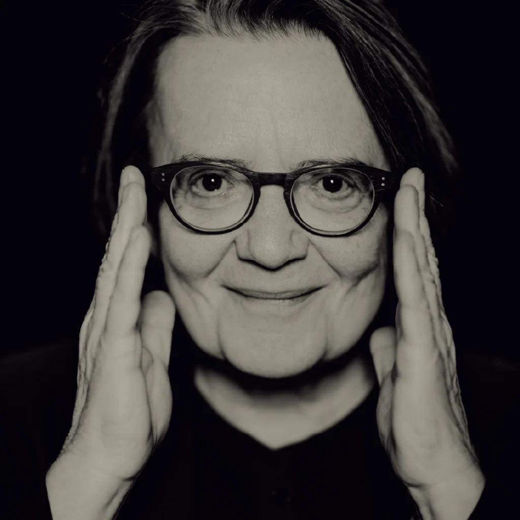 Primo piano in bianco e nero della regista Agnieszka Holland, con le mani che incorniciano il viso e un sorriso enigmatico.