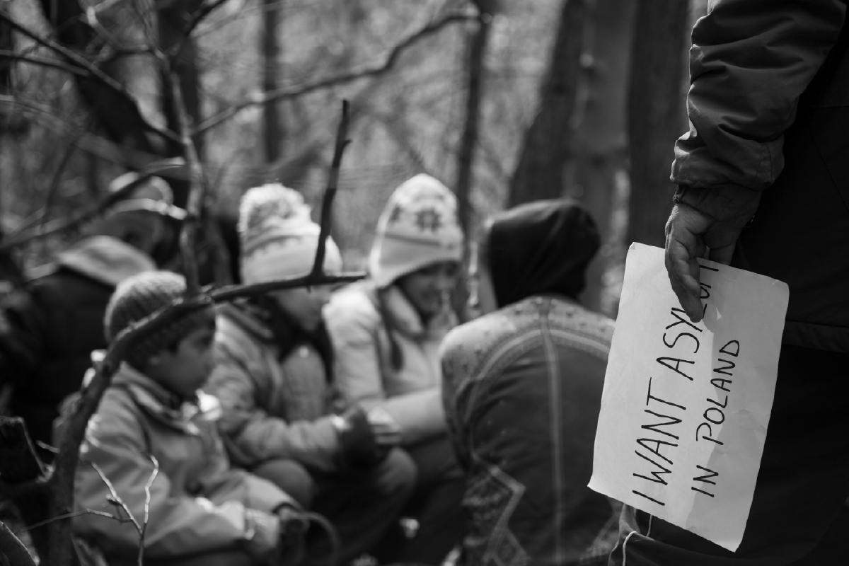 Primo piano su una mano che regge un foglio con la scritta “I want asylum in Poland”, con bambini seduti sullo sfondo nel bosco.