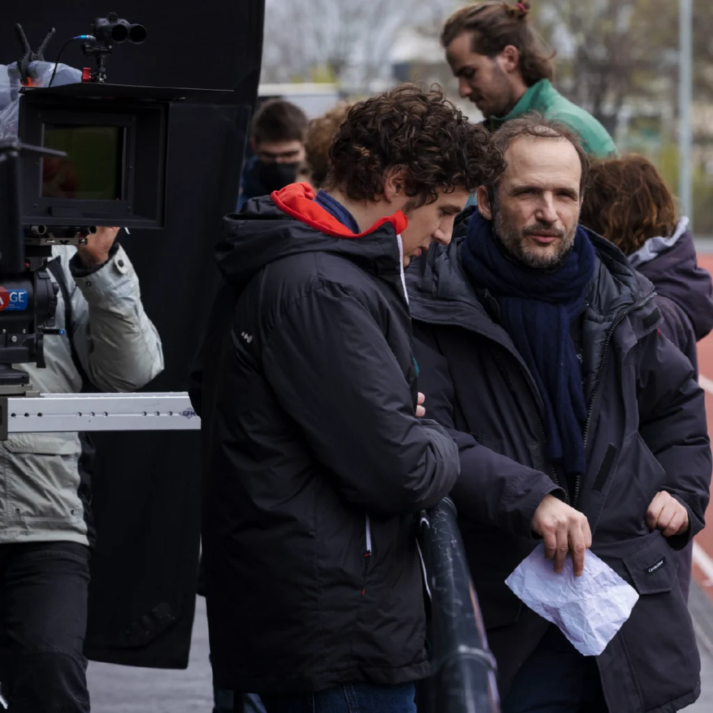 Il regista Thomas Lilti sul set di “Guida pratica per insegnanti”, mentre discute una scena con l’attore Vincent Lacoste durante le riprese, entrambi vestiti in abiti invernali.